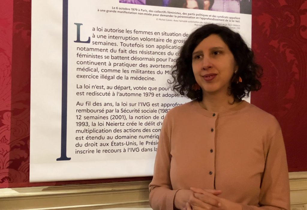 Photo de BIbia Pavard posant devant un panneau de l'exposition Loi sur l'IVG 1974, aux Archives nationales en mars 2024.